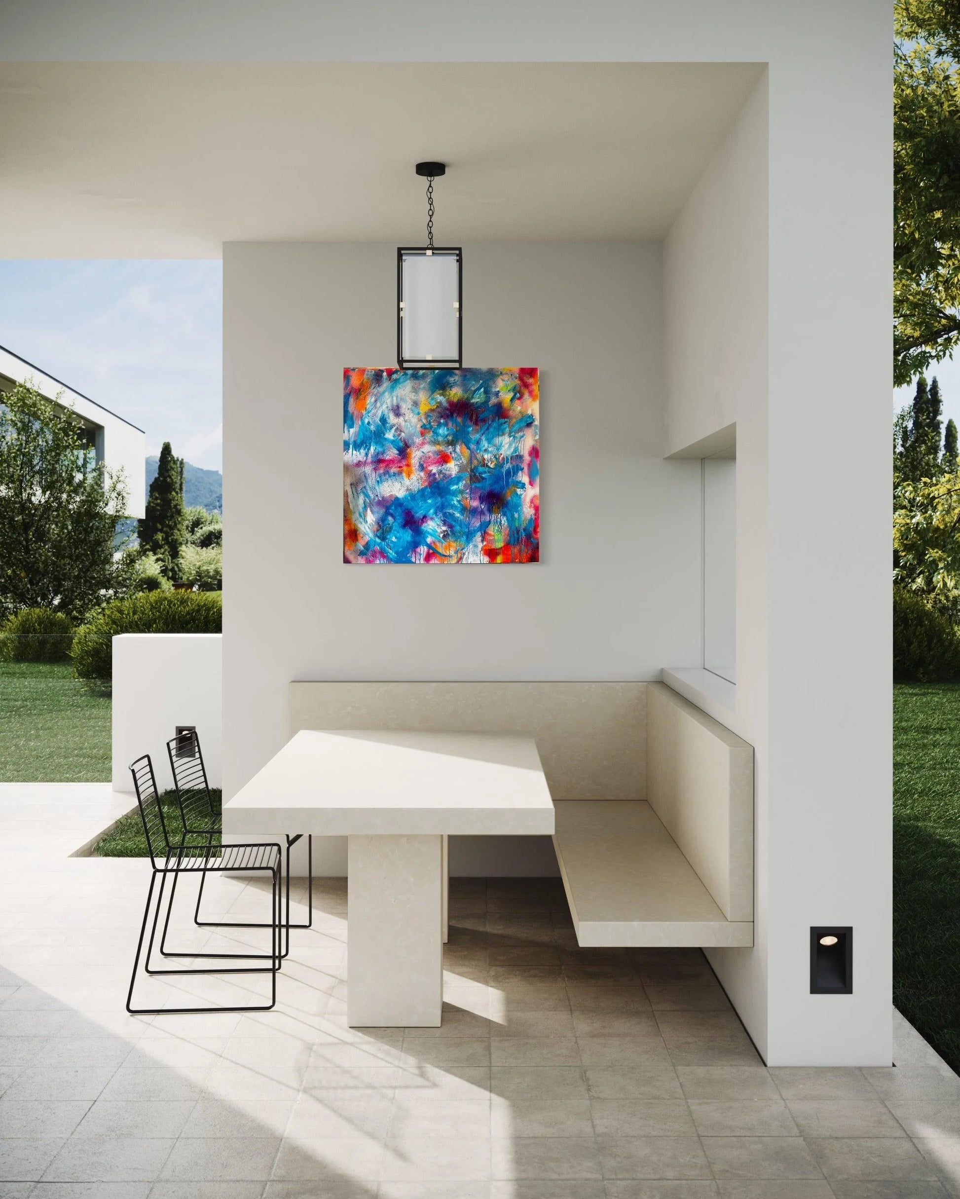 Shown is a covered patio with fixed bench seating and stone table, two additional black wire chairs complete the set. An outdoor light hangs from the centre and the painting can be seen on the wall in front. a neatly mown lawn can be viewed either side of the stone patio floor.