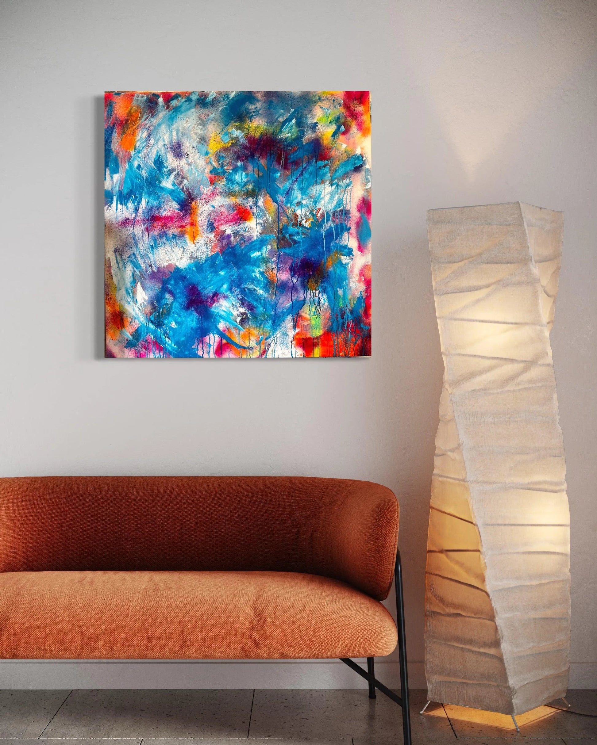 The artwork is hung on a white wall above a burnt orange sofa that has black metal legs, an elaborately twisted tall paper lamp is beside the sofa.