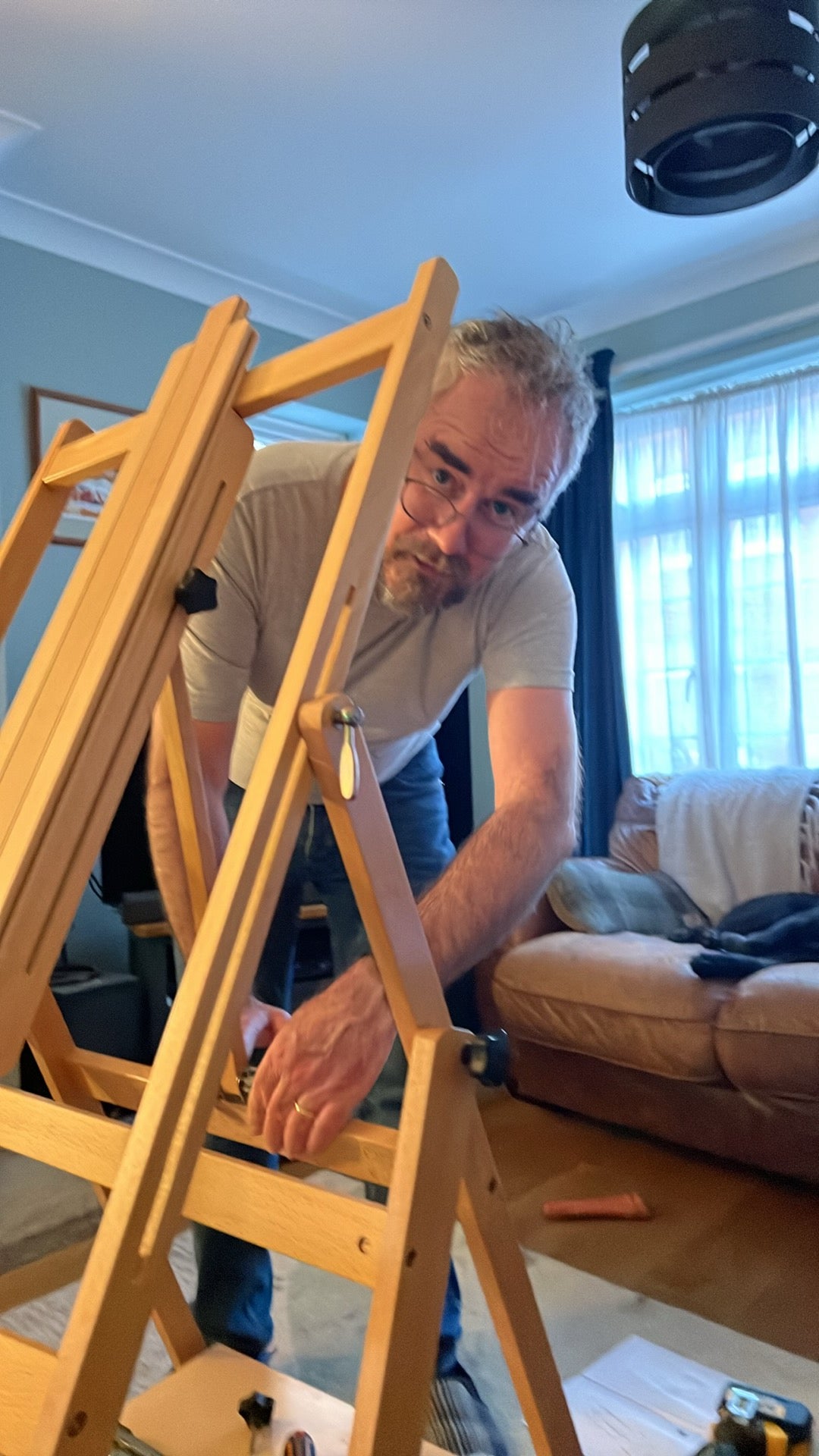 A man can be seen putting together a wooden easel. He is wearing jeans and a grey T-shirt, he has glasses, grey hair and a goatee.  In the background is a leather settee.