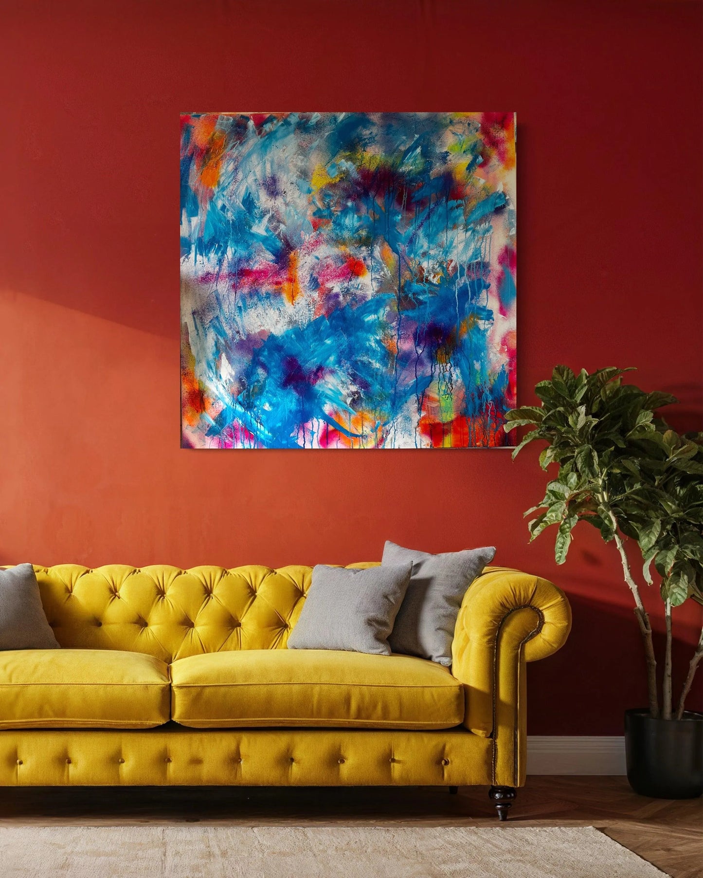 The artwork is hung on a deep orange wall above a yellow Chesterfield style sofa, with a cream rug on the floor in front of the sofa and a large pot plant to the right. There are also taupe scatter cushions on the sofa, one on the left and two on the right.