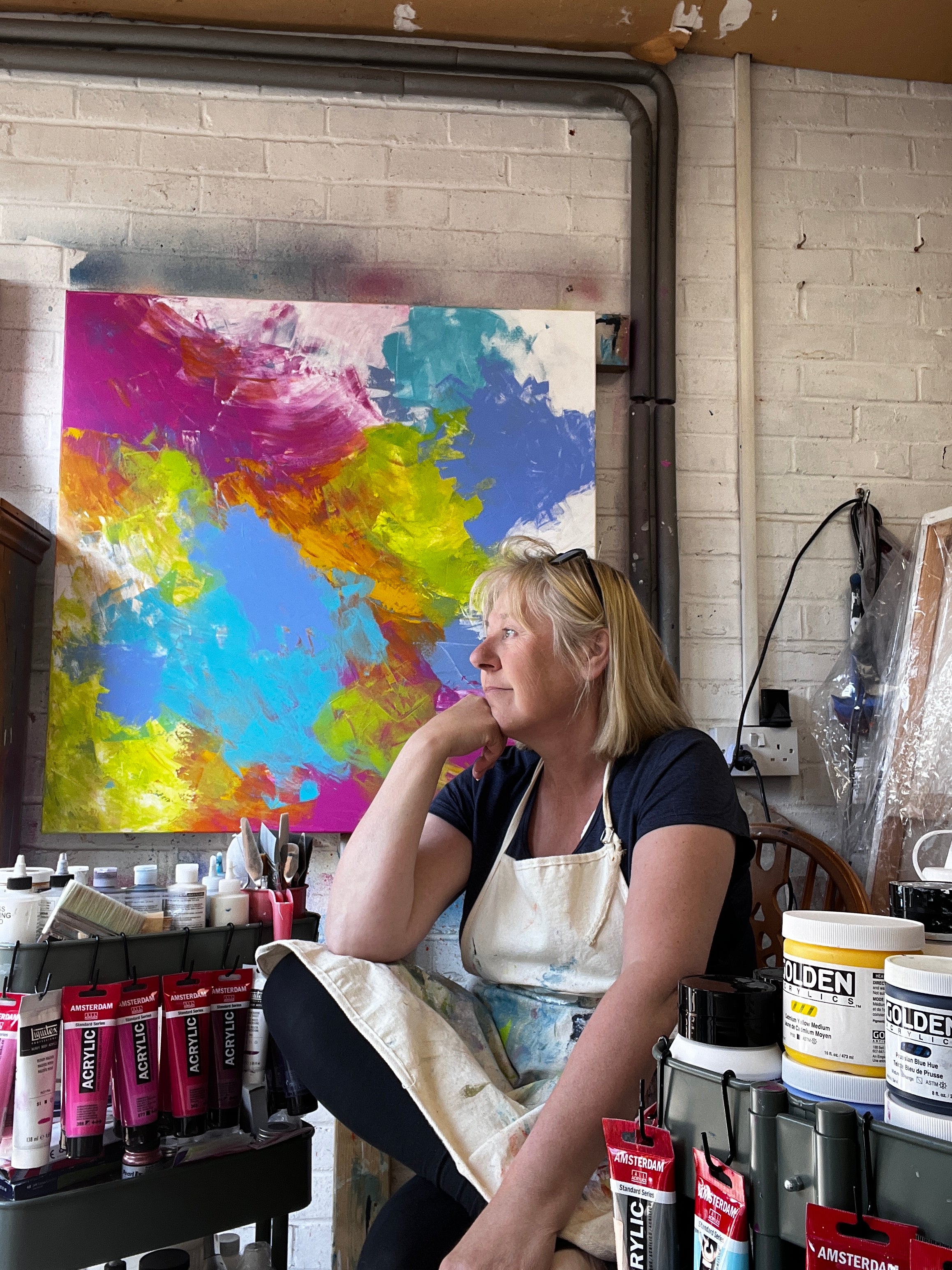 Inside my garage studio, a lady with blonde hair and glasses on her head, sits in the centre of the photograph. Looking out to the left of the photograph thoughtfully resting her chin on her hand, legs crossed. Sitting amongst pain ing paraphinalia, paints hanging from tiered trolleys. A large colourful canvas is on the wall in the background.