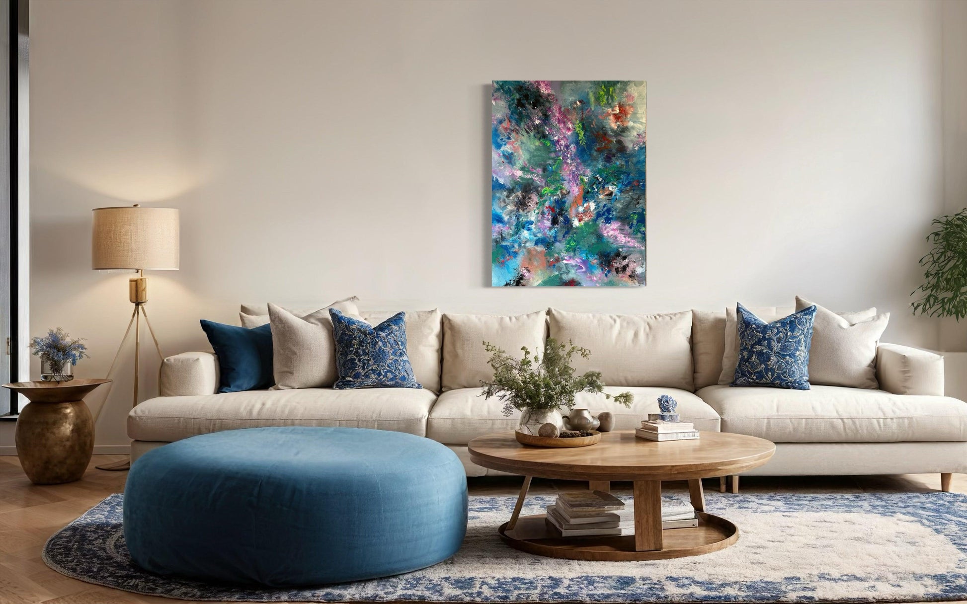 A living room showing the artork above a long cream sofa with blue and cream scatter cushions, a round wooden coffee table is in the foreground with a wooden tray holding a plant aand ephemaera, beside a pile of 3 books. A are circular blue pouffe stands to the left of the table also in front of the sofa. A standard lamp with cream shade and brass side table lie to the left of the photogrph with a plant to the right. The floor is parquet with a large blue and cream rug.